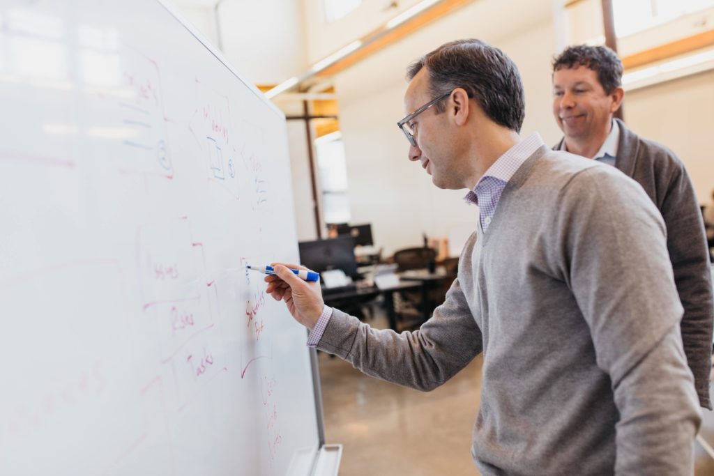 Don Stavneak writes on a whiteboard while Rod Smith smiles and watches.